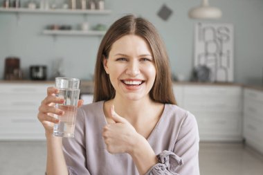 Elinde bir bardak su olan genç ve güzel bir kadın evde baş parmağını kaldırıyor.