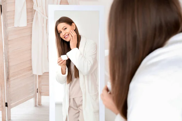 Beautiful young woman applying cream near mirror