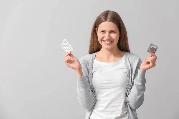 Young woman with birth control pills and condom on grey background