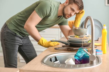 Mutfakta kirli bulaşık yığınına bakan hoşnutsuz bir adam.