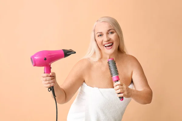 Beautiful mature woman with hair dryer and brush singing on color background