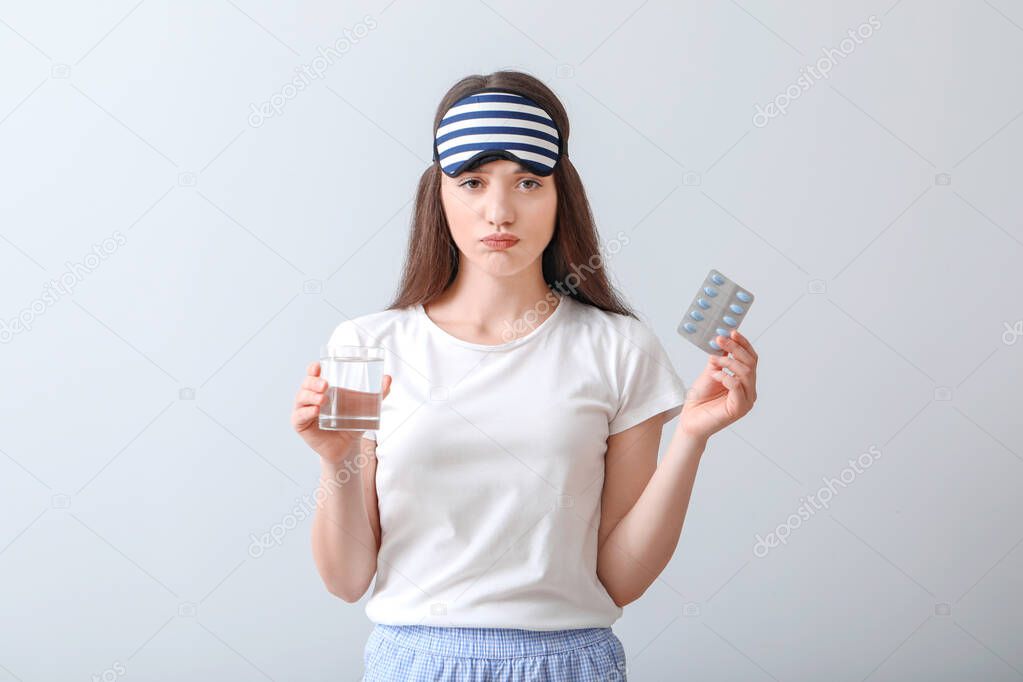 Sad woman with sleeping pills and glass of water on light background