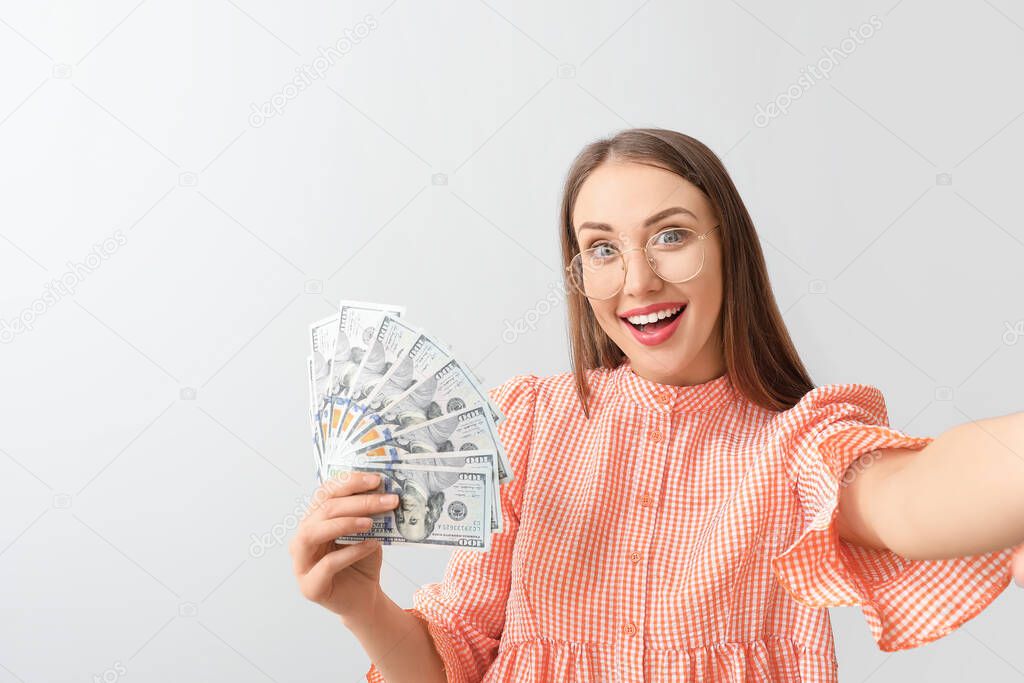 Happy young woman taking selfie with money on light background