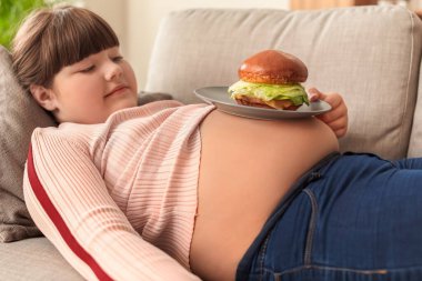 Evdeki kanepede hamburger yiyen şişman bir kız.
