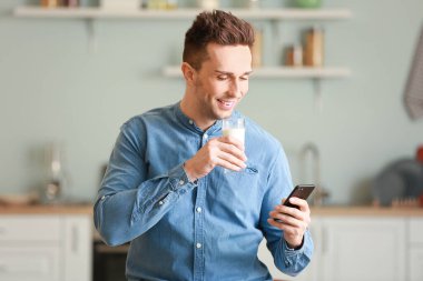 Cep telefonlu genç adam mutfakta süt içiyor.