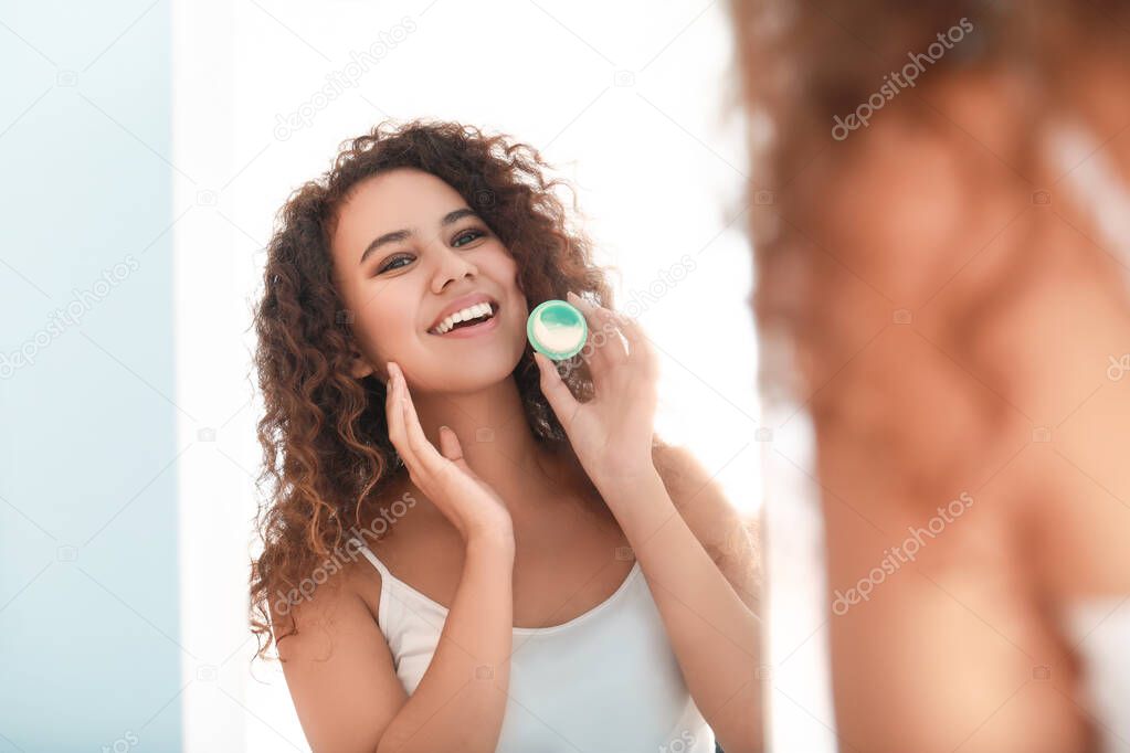 Beautiful young African-American woman applying cream at home