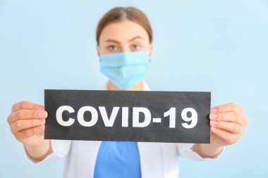 Koruyucu maskeli bir doktor elinde renkli arka planda COVID-19 yazılı kağıt çarşaf tutuyor. Salgın kavramı