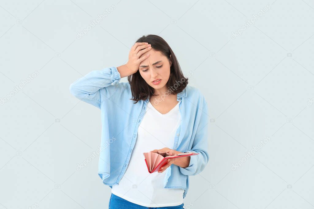 Worried young woman with empty wallet on light background