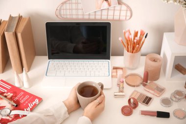 Makyaj sanatçısı modern laptop ve kozmetik ürünleri ile masada çay içiyor.