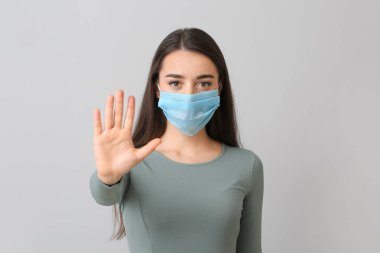 Tıbbi maskeli genç kadın gri arka planda DUR hareketi gösteriyor.