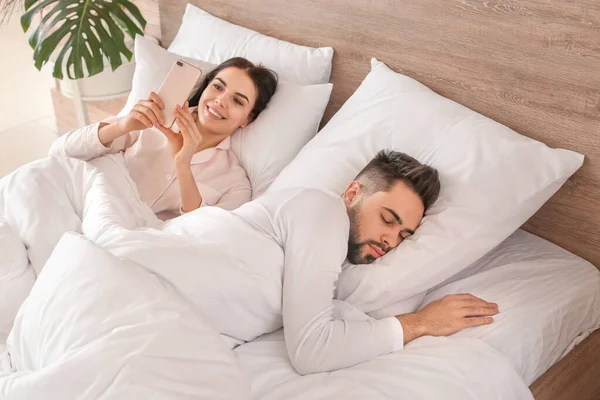 夫がベッドで寝ている間に携帯電話を使う若い女性 — ストック写真