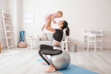 Young sporty mother and her baby doing exercises with fitball at home clipart