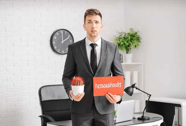 Young businessman holding cactus and paper with text HEMORRHOIDS in office