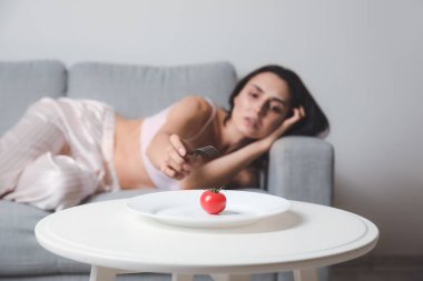 Young woman suffering from anorexia and plate with tomato on table clipart