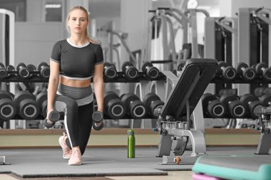 Spor salonunda dambıllarla spor yapan genç bir bayan.