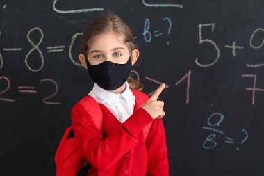 Okul tahtasının yanında maske takan küçük bir kız.