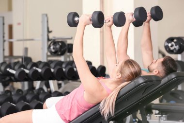 Spor salonunda dambıllarla spor yapan genç bir çift.