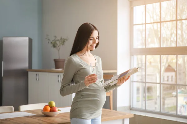 Beautiful pregnant woman with magazine and glass of water in kitchen