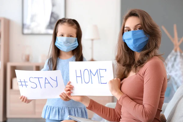 Mother and little daughter in protective masks holding papers with text STAY HOME at home