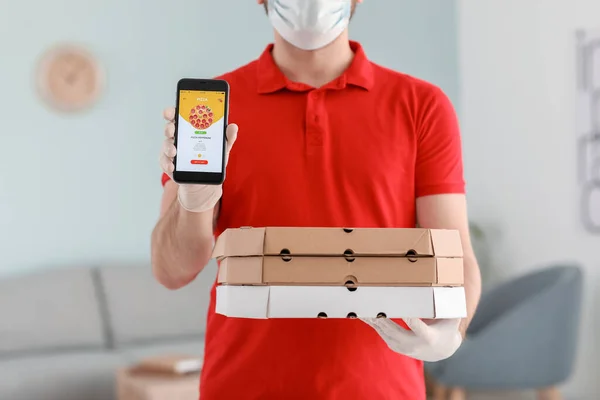 Male courier of food delivery company in medical mask and with mobile phone indoors