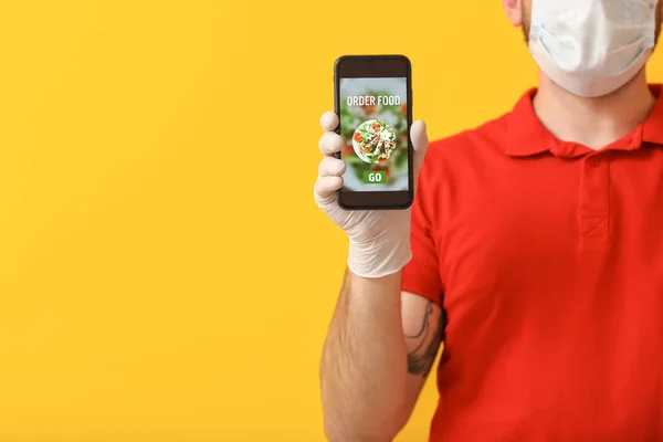 Male courier of food delivery company in medical mask and with mobile phone on color background