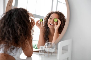 Beautiful young African-American woman with cucumber slices in bathroom clipart