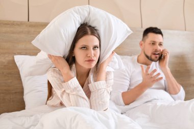 Young woman cannot sleep because of her husband talking by mobile phone in bedroom clipart