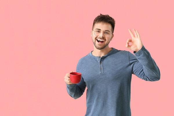 Young man with cup of hot coffee showing OK on color background
