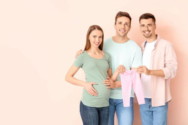 Schwules Paar Und Schwangere Frau Mit Babykleidung Auf Farbigem Hintergrund — Stockfoto