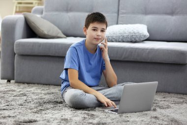Evde dizüstü bilgisayar ve cep telefonu kullanan küçük bir çocuk.