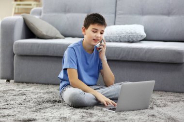 Evde dizüstü bilgisayar ve cep telefonu kullanan küçük bir çocuk.