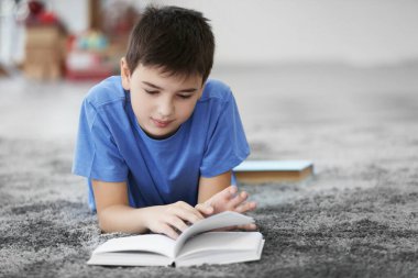 Küçük çocuk evde yerde kitap okuyor.