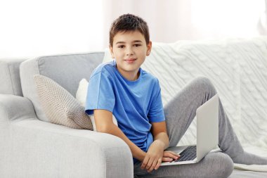 Evdeki kanepede oturan dizüstü bilgisayarı olan çocuk.