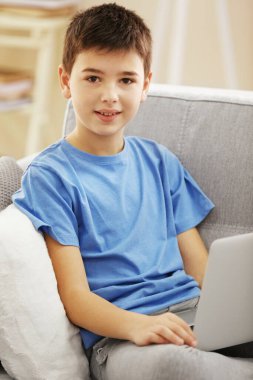 Evdeki kanepede oturan dizüstü bilgisayarı olan çocuk.