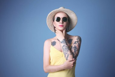 Mavi arka planda şapka ve güneş gözlüğü takan dövmeli güzel bir genç kadın.
