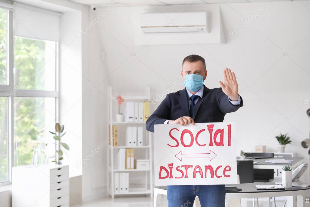 Mature man holding paper with text SOCIAL DISTANCE in office. Concept of epidemic
