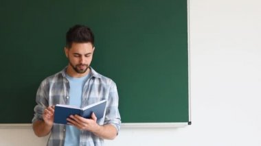 Sınıfta tahtanın yanında kitap okuyan bir erkek öğrenci.