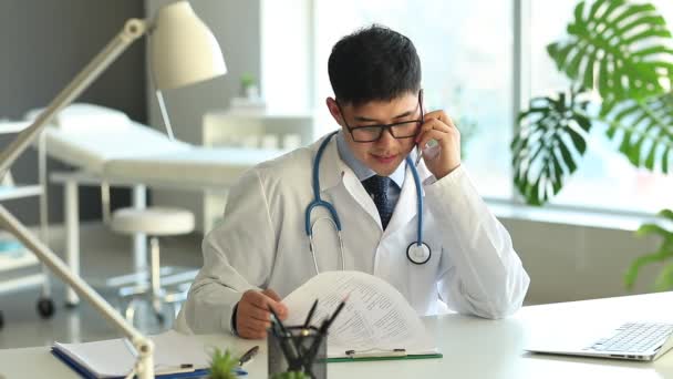 Ασιάτης Γιατρός Μιλάει Τηλεφωνικά Στο Τραπέζι Στην Κλινική — Αρχείο Βίντεο