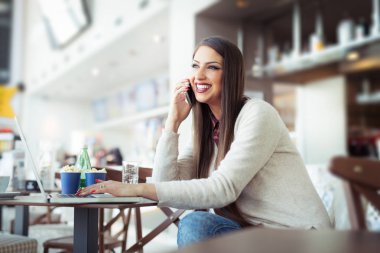 Dizüstü bilgisayar ile kafeteryada oturan ve cep telefonuyla genç kadın