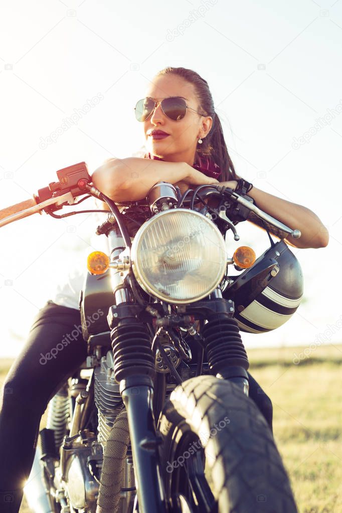 Outdoor lifestyle portrait of young biker woman sitting on a vintage custom motorcycle
