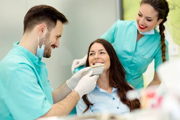 Dentist checking and selecting color of young woman's teeth — Stock Photo, Image