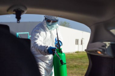 Koruyucu giysili, maskeli bir adam arabanın içini dezenfekte ediyor..