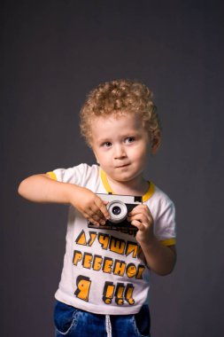 Gri bir arka plan - studio fotoğraf üzerinde bir kamera küçük kıvırcık saçlı çocuk. Fotoğraf çekmek çocuk öğrenir. 