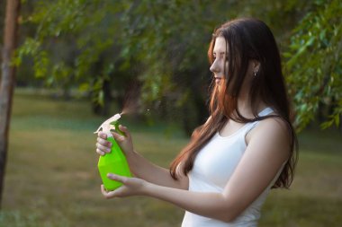 Young woman spraying water on herself from a spray bottle in a summer park.