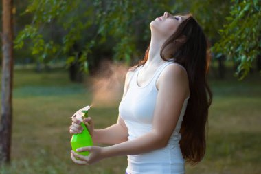 Young woman spraying water on herself from a spray bottle in a summer park.