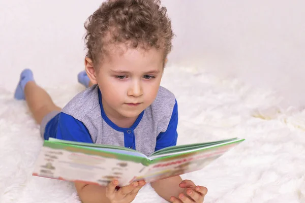 小さな男の子がふわふわのベッドの上に横たわって本を読んでいます 子供たちに読書 写真を教える — ストック写真