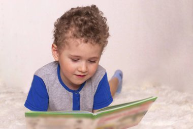 Küçük bir çocuk kabarık bir yatak örtüsüne uzanıp kitap okuyor. Çocuklara okumayı öğretmek, bir fotoğraf.