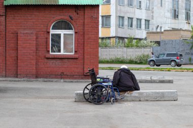 Boş tekerlekli sandalye, bir kadın beton bir levhada oturuyor, arkadan görüşü var. Yoksulluk ve hastalık kavramı.