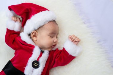 Kırmızı Noel Baba kostümü giyen bebek beyaz kürk halıda uyuyor. Noel ve Yeni Yıl tatili kutlaması kavramı.