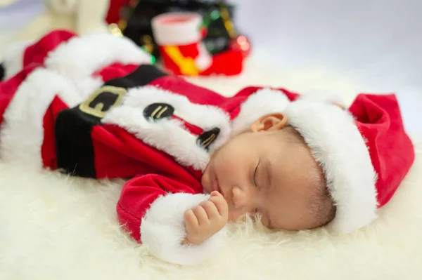 Kırmızı Noel Baba kostümü giyen bebek beyaz kürk halıda uyuyor. Noel ve Yeni Yıl tatili kutlaması kavramı.